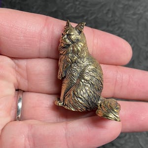 May include: A detailed, golden-colored cat figurine. The cat is sitting upright with its tail curled. The figurine has textured fur and is held in a person's hand. The cat's features are well-defined, and the overall design is intricate.