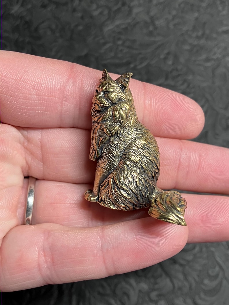 May include: A detailed, golden-colored cat figurine. The cat is sitting upright with its tail curled. The figurine has textured fur and is held in a person's hand. The cat's features are well-defined, and the overall design is intricate.