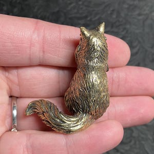 May include: A detailed, golden-colored fox figurine. The fox is in a sitting position with its tail curled. The figurine has textured fur and is held in a person's hand. The fox is approximately 1.5 inches tall.
