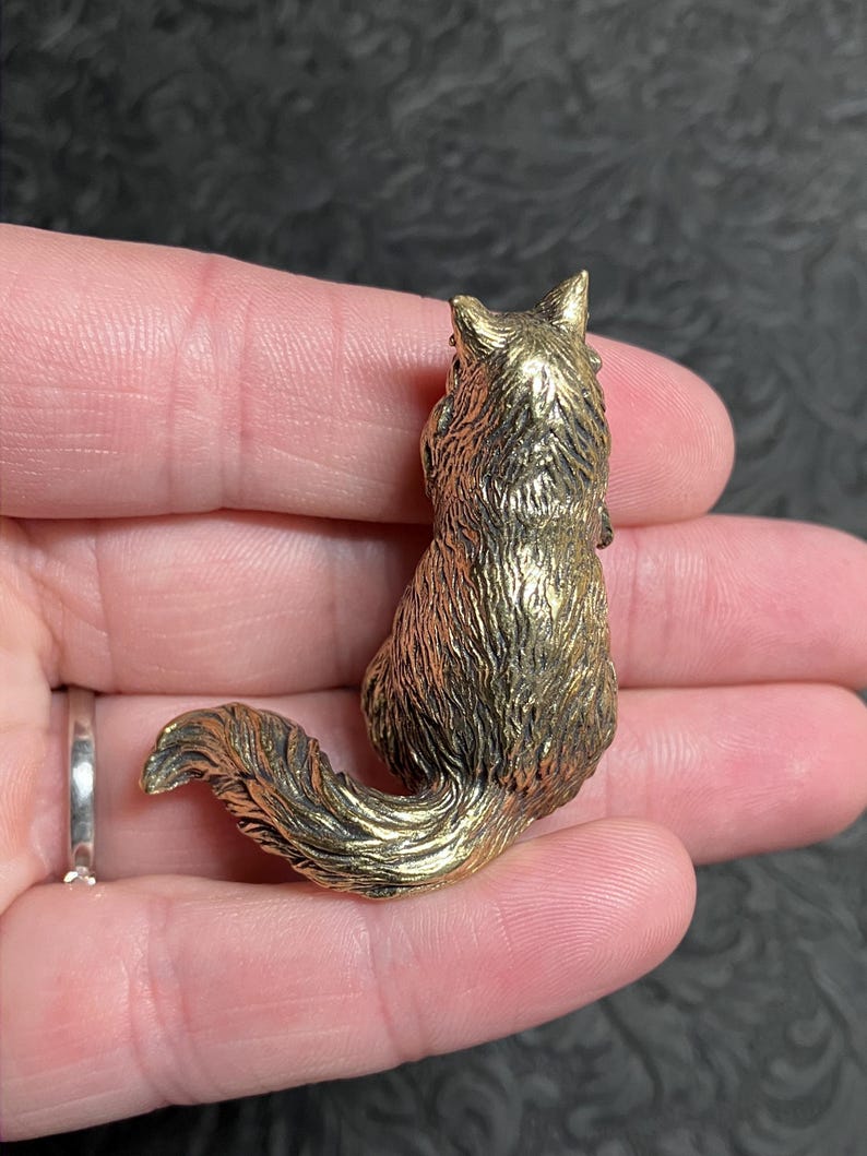 May include: A detailed, golden-colored fox figurine. The fox is in a sitting position with its tail curled. The figurine has textured fur and is held in a person's hand. The fox is approximately 1.5 inches tall.