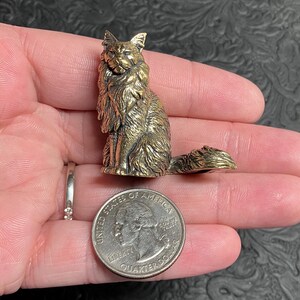 May include: A detailed brass cat figurine, seated with a long tail, held in a hand next to a quarter for scale. The cat has detailed fur and a regal pose. The quarter is 0.955 inches in diameter.