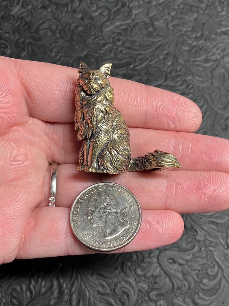 May include: A detailed brass cat figurine, seated with a long tail, held in a hand next to a quarter for scale. The cat has detailed fur and a regal pose. The quarter is 0.955 inches in diameter.