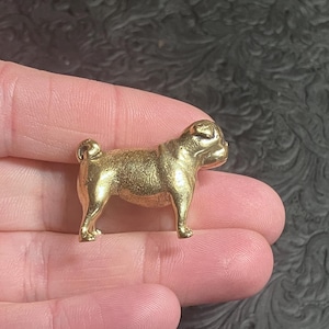 May include: A small, gold-colored pug dog figurine. The pug is standing with its tail curled and its features are detailed. The figurine is held in a person's hand, and the background is a dark, textured surface.