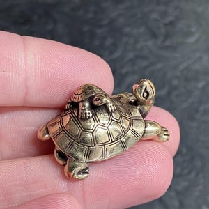 May include: A small, detailed brass turtle figurine with a smaller turtle on its back. The figurine has a textured shell and is held between fingers against a dark background. The turtle is a decorative item.