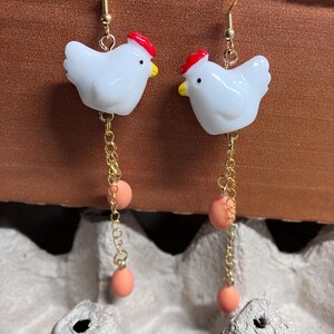 Pode incluir: Um par de brincos de ouro pendentes com pingentes de frango de cer&acirc;mica branca com cristas vermelhas e uma corrente pendente com tr&ecirc;s contas cor de p&ecirc;ssego.