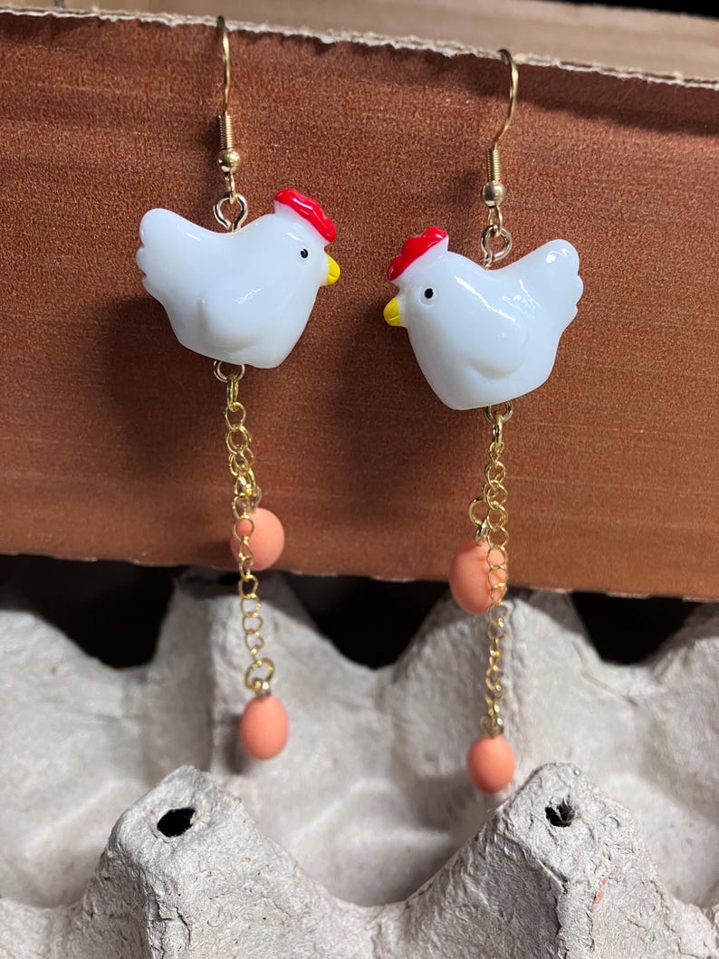 Pode incluir: Um par de brincos de ouro pendentes com pingentes de frango de cer&acirc;mica branca com cristas vermelhas e uma corrente pendente com tr&ecirc;s contas cor de p&ecirc;ssego.