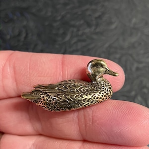 Pode incluir: Pequena e detalhada estatueta de pato em latão. O pato tem penas texturizadas e um bico detalhado. A estatueta é segurada na mão de uma pessoa, mostrando seu tamanho e artesanato. O pato é um objeto decorativo.