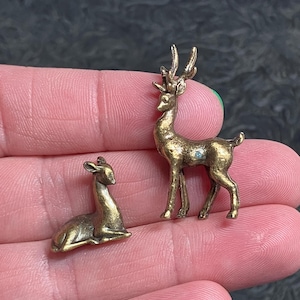 May include: Two small, antique-style brass deer figurines. One deer is standing, the other is lying down. The standing deer has detailed antlers and a small blue gem. These miniature sculptures are likely decorative.