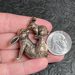 Pode incluir: Uma estatueta de metal em tom de bronze apresentando dois coelhos, um coelho maior sentado e um coelho menor empoleirado nas costas, posicionada ao lado de uma moeda de 25 centavos dos EUA de 2023 para escala, com textura de pelo detalhada.