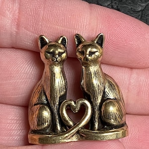 Pode incluir: Pequena estatueta dourada de dois gatos sentados lado a lado, com as caudas formando um coração. Os gatos têm detalhes finos e estão sobre uma pequena base. A estatueta é segurada em uma mão.
