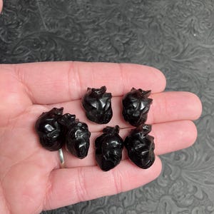 May include: Seven small, black, heart-shaped carvings are displayed in a person's open hand. The carvings have a glossy finish and are arranged in a cluster. The background is a textured, dark surface.