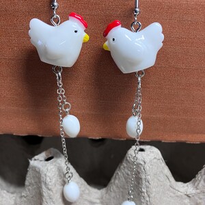 Pode incluir: Brincos de cer&acirc;mica branca em forma de frango com cristas vermelhas e contas brancas penduradas em correntes prateadas.