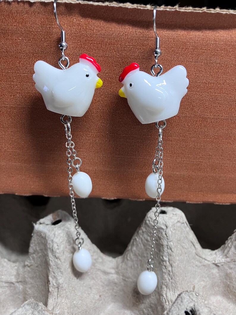 Pode incluir: Brincos de cer&acirc;mica branca em forma de frango com cristas vermelhas e contas brancas penduradas em correntes prateadas.