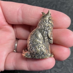 May include: A small, bronze-colored figurine of a seated dog with detailed fur texture. The dog has pointed ears and a long, flowing coat. The figurine is approximately 1.5 inches tall and has a vintage appearance.