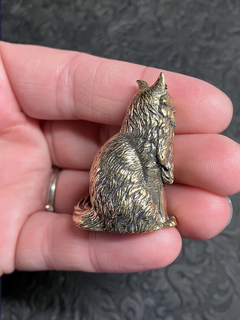 May include: A small, bronze-colored figurine of a seated dog with detailed fur texture. The dog has pointed ears and a long, flowing coat. The figurine is approximately 1.5 inches tall and has a vintage appearance.