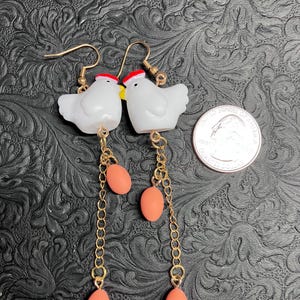 Pode incluir: Um par de brincos pendentes com galinhas de cer&acirc;mica branca com cristas vermelhas e bicos amarelos. Cada galinha est&aacute; presa a uma corrente dourada com tr&ecirc;s contas de cor coral em forma de ovo.