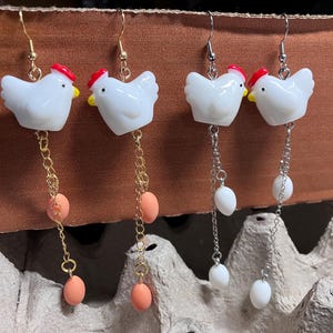 Pode incluir: Um par de brincos pendentes com pingentes de frango de cer&acirc;mica branca com detalhes vermelhos e amarelos. Os pingentes s&atilde;o presos a correntes douradas com tr&ecirc;s contas cor de p&ecirc;ssego em cada corrente.