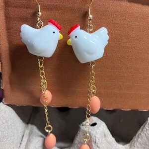 Pode incluir: Um par de brincos de ouro pendurados com pingentes de frango de cer&acirc;mica branca com cristas vermelhas e tr&ecirc;s contas de ovo cor de p&ecirc;ssego em cada brinco.