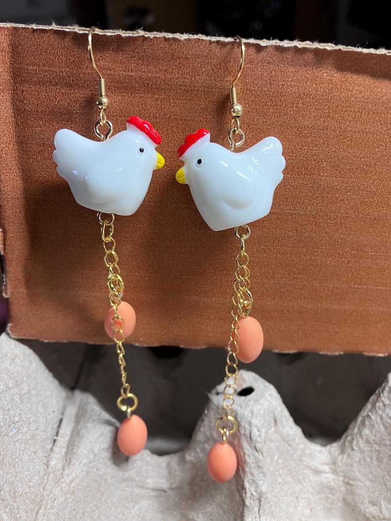Pode incluir: Um par de brincos de ouro pendurados com pingentes de frango de cer&acirc;mica branca com cristas vermelhas e tr&ecirc;s contas de ovo cor de p&ecirc;ssego em cada brinco.