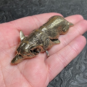 May include: A detailed, golden-colored metal fox figurine in a resting pose. The fox has pointed ears, a long snout, and textured fur detailing. The figurine is lying on a person's open palm, showcasing its size and craftsmanship.