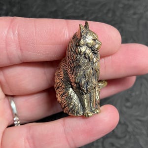 May include: A detailed, golden-colored cat figurine. The cat is sitting upright with its fur meticulously sculpted. The figurine is held in a hand, showcasing its size and intricate design. The cat's features are clearly defined, giving it a lifelike appearance.