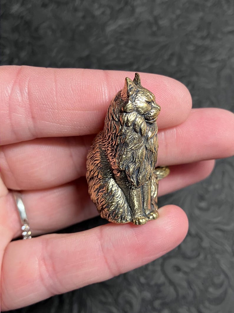 May include: A detailed, golden-colored cat figurine. The cat is sitting upright with its fur meticulously sculpted. The figurine is held in a hand, showcasing its size and intricate design. The cat's features are clearly defined, giving it a lifelike appearance.