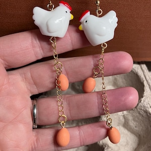 Pode incluir: Um par de brincos pendentes com pingentes de frango de cer&acirc;mica branca com cristas e bicos vermelhos. Cada pingente &eacute; preso a uma corrente dourada com tr&ecirc;s contas de ovo cor de p&ecirc;ssego.