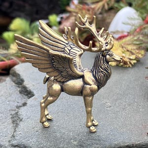 May include: A detailed brass figurine of a winged deer, also known as a stag. The sculpture features intricate details, including antlers, feathered wings, and a muscular body. The deer is standing on a gray stone surface, with greenery in the background.