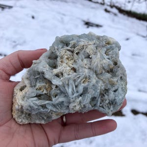 May include: A light blue mineral specimen with a crystalline structure, held in a hand. The rock has a rough, textured surface with areas of white and tan coloration. The background shows a snowy landscape.