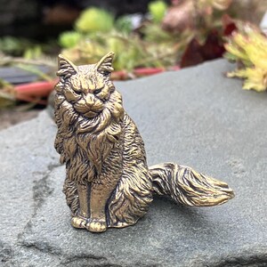 May include: A detailed brass figurine of a cat in a seated position. The cat has a long, flowing mane and tail, with an alert expression. The figurine is a golden color and sits on a gray stone surface.