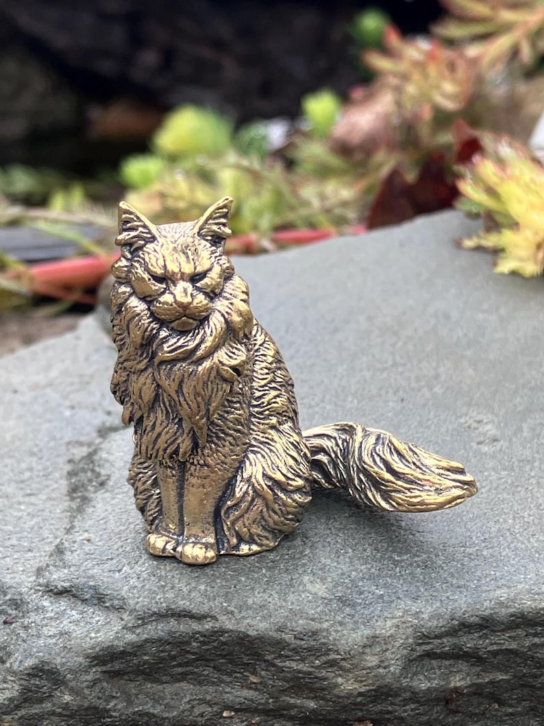 May include: A detailed brass figurine of a cat in a seated position. The cat has a long, flowing mane and tail, with an alert expression. The figurine is a golden color and sits on a gray stone surface.