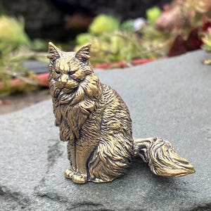 May include: A detailed brass cat figurine, posed in a seated position with its tail curled. The sculpture has a textured, fur-like appearance and is set against a blurred background of greenery and a gray stone surface.