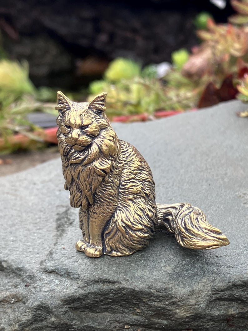 May include: A detailed brass cat figurine, posed in a seated position with its tail curled. The sculpture has a textured, fur-like appearance and is set against a blurred background of greenery and a gray stone surface.