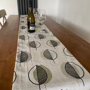 May include: A white linen table runner with a black and green leaf pattern. The runner is on a wooden table with a bottle of wine and two wine glasses.