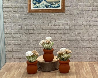 May include: Three small terracotta pots with white and tan flowers sit on a wooden surface. A framed print of a Japanese wave painting hangs on a brick wall in the background. The pots are arranged in a decorative display.