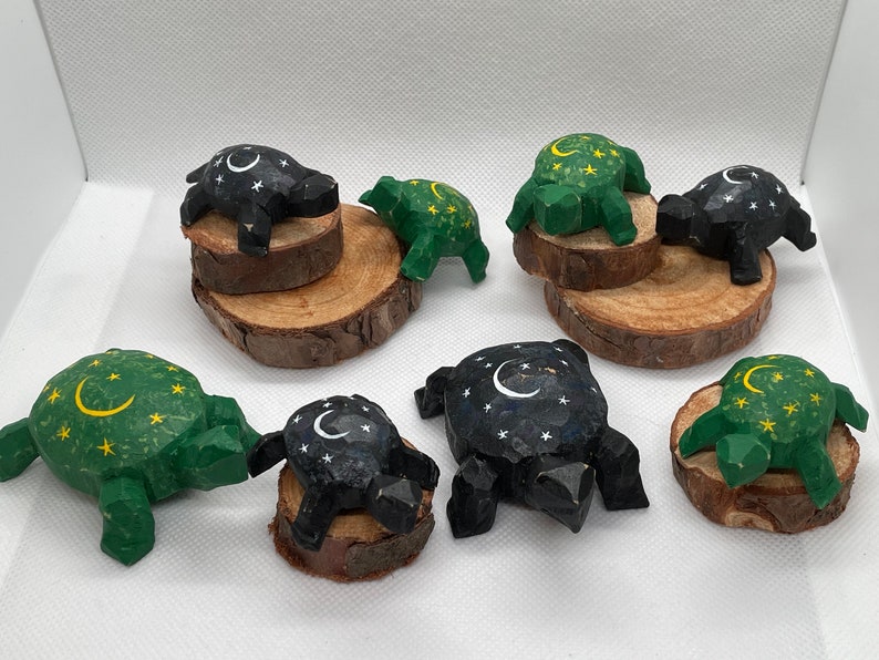 K&ouml;nnte beinhalten: Sieben h&ouml;lzerne Schildkr&ouml;tenfiguren mit einem Halbmond- und Sternenmuster. Die Schildkr&ouml;ten sind in Gr&uuml;n- und Schwarzt&ouml;nen bemalt und auf kleinen Holzscheiben ausgestellt.