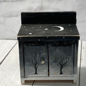 May include: A small black wooden cabinet with two doors. The doors are painted with a black and white tree design. The top of the cabinet is painted black with a white crescent moon and stars.