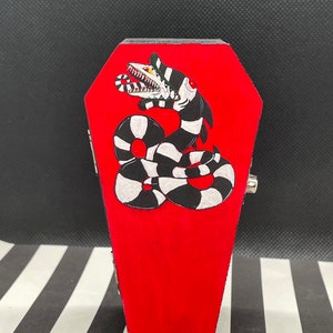 May include: A red wooden coffin with a black and white snake design. The snake has a large mouth with sharp teeth and is coiled around itself. The coffin is a small decorative box.