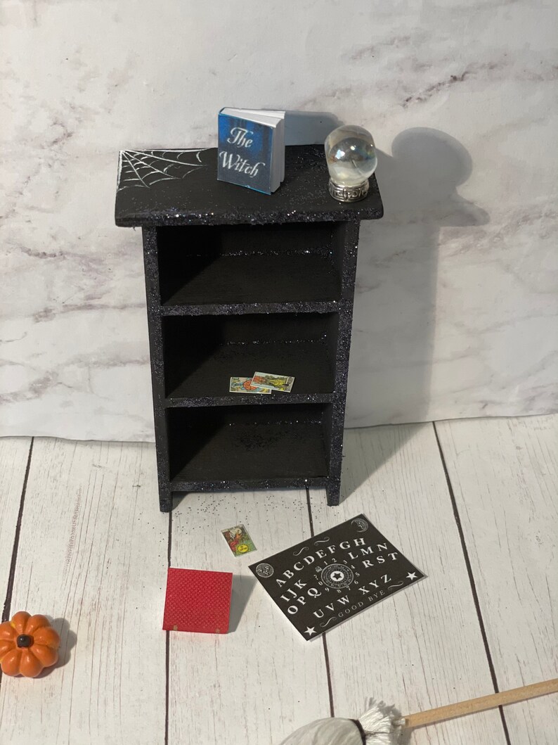 May include: Black bookshelf with four shelves, a spiderweb design on the top shelf, a book titled "The Witch", a crystal ball, and a Ouija board on the floor.
