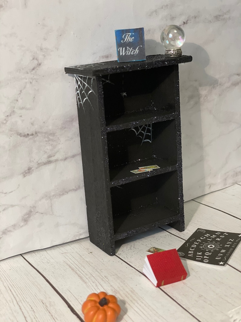 May include: A black wooden shelf with three shelves, decorated with glitter and spiderwebs. The shelf is topped with a crystal ball and a sign that reads "The Witch".