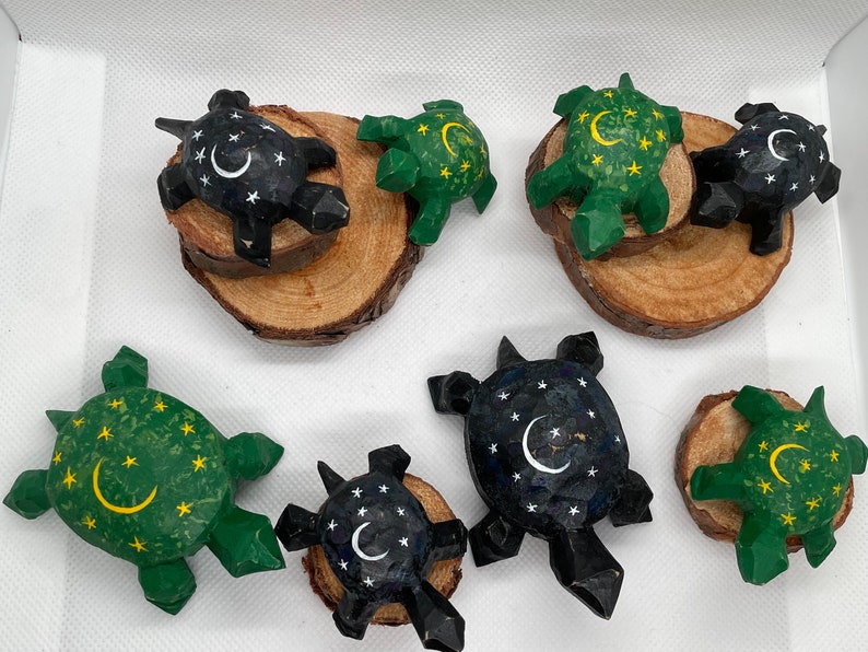 K&ouml;nnte beinhalten: Eine Sammlung von acht handgeschnitzten Holz-Schildkr&ouml;ten, jede mit einem anderen Design bemalt. Einige Schildkr&ouml;ten haben eine gr&uuml;ne Schale mit gelben Sternen und einem Halbmond, w&auml;hrend andere eine schwarze Schale mit wei&szlig;en Sternen und einem Halbmond haben.
