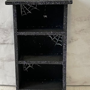 May include: Black wooden shelf with three shelves, decorated with glitter and white spiderwebs.