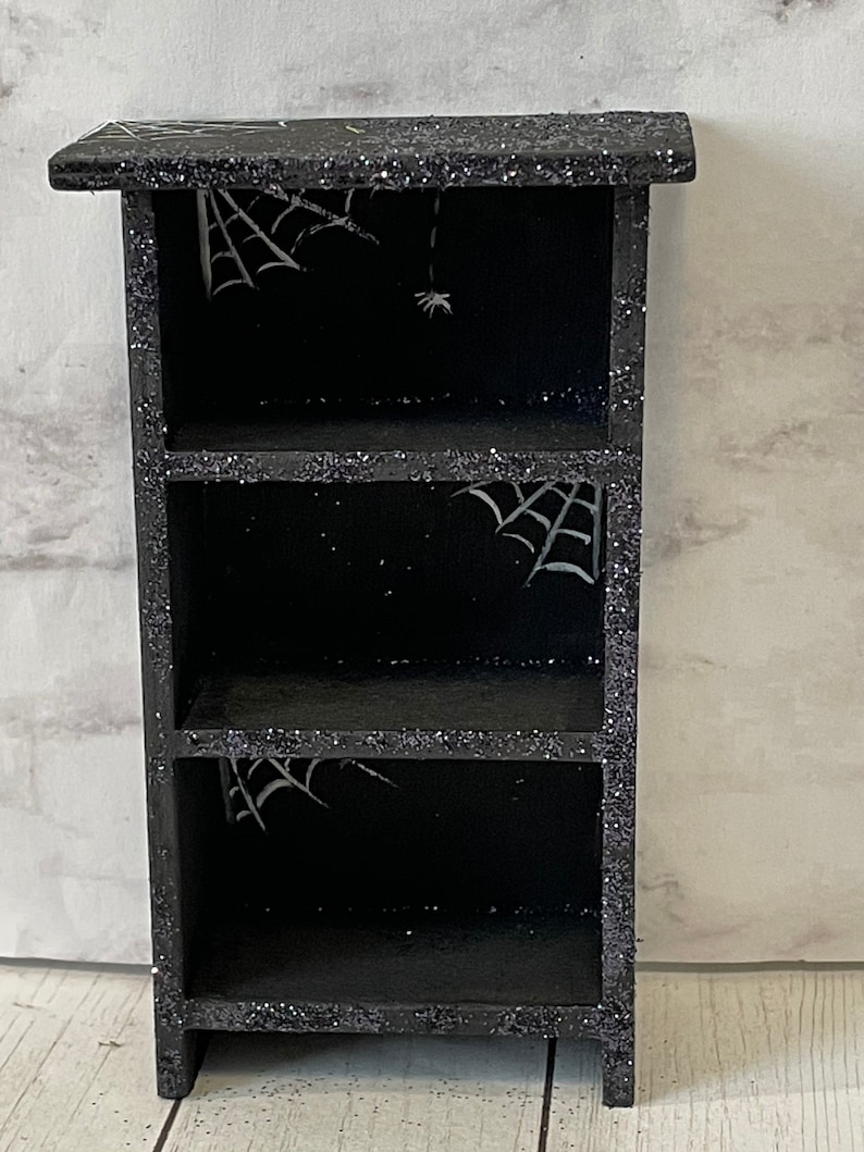 May include: Black wooden shelf with three shelves, decorated with glitter and white spiderwebs.