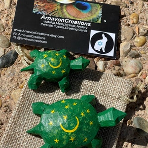K&ouml;nnte beinhalten: Zwei h&ouml;lzerne Schildkr&ouml;tenfiguren, die gr&uuml;n mit gelben Sternen und einem Halbmond bemalt sind. Die Schildkr&ouml;ten stehen auf einer braunen Jutestoff-Oberfl&auml;che.