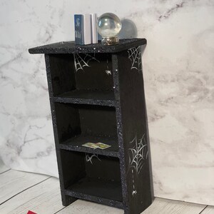 May include: A black wooden bookshelf with three shelves, decorated with white spiderwebs and glitter. The bookshelf has a small crystal ball and a book on top. There is a red book and a Ouija board on the floor in front of the bookshelf.
