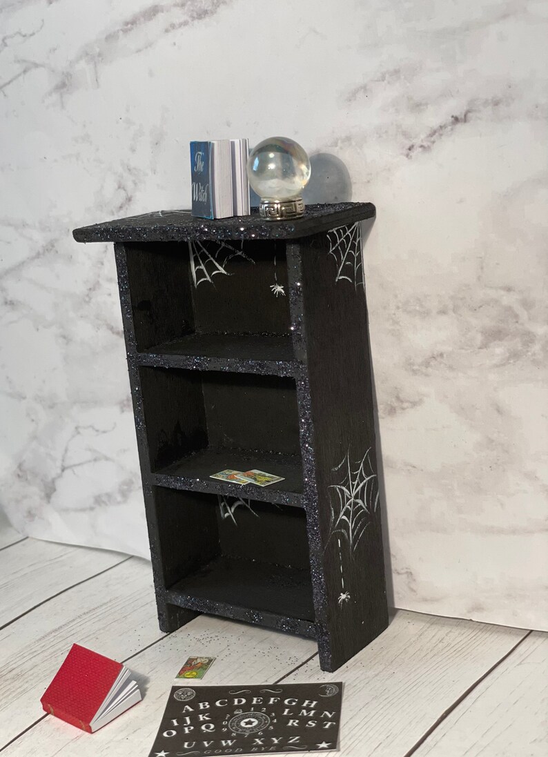 May include: A black wooden bookshelf with three shelves, decorated with white spiderwebs and glitter. The bookshelf has a small crystal ball and a book on top. There is a red book and a Ouija board on the floor in front of the bookshelf.