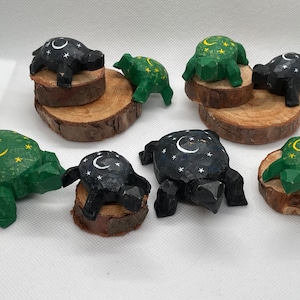 May include: Eight wooden turtle figurines, painted in black and green with a crescent moon and stars design. The turtles are displayed on small wooden rounds.