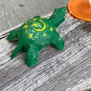 K&ouml;nnte beinhalten: Eine kleine gr&uuml;ne Holz-Schildkr&ouml;te mit einem gelben Halbmond und Sternen, die auf ihren Panzer gemalt sind. Die Schildkr&ouml;te sitzt auf einem St&uuml;ck verwittertem Holz.