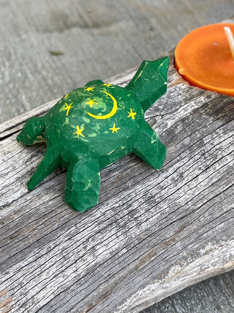 K&ouml;nnte beinhalten: Eine kleine gr&uuml;ne Holz-Schildkr&ouml;te mit einem gelben Halbmond und Sternen, die auf ihren Panzer gemalt sind. Die Schildkr&ouml;te sitzt auf einem St&uuml;ck verwittertem Holz.