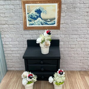 May include: Miniature scene featuring a black nightstand with three drawers, topped with a small potted arrangement of white flowers and a ladybug. Two additional flower pots with ladybugs and white flowers sit in front. A framed print of a wave scene hangs on the brick wall.